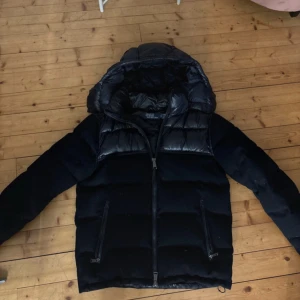 Svart pufferjacka Polo Ralph Lauren - Snygg svart pufferjacka från Polo Ralph Lauren med glansiga partier upptill och på huvan. Jackan har dragkedja framtill, två dragkedjefickor och en rejäl huva. Perfekt för kalla dagar och riktigt stilren look. Inga skavanker eller skador på den alls!  Kom med bud! 