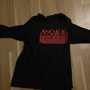 Svart långärmad tröja Hugo Boss - Svart långärmad tröja från Hugo Boss med stort rött BOSS-tryck på bröstet. Tröjan har en klassisk passform och är tillverkad i mjuk bomull som känns skön mot huden. Perfekt för dig som gillar stilrena logotröjor med attityd.