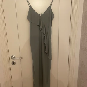 Ljusgrön medel långklänning med volang - Säljer en elegant ljusgrön långklänning från H&M med smala axelband och snygg volangdetalj framtill. Klänningen har en figurnära passform och är tillverkad i ett lätt, mjukt polyestertyg som faller fint. 