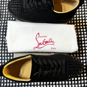 Christian Louboutin svarta sneakers med nitar - Svarta sneakers från Christian Louboutin i mocka med svarta nitar på tån och klassisk röd sula. Skorna har snörning, rund tå och beige innersida. Perfekta för dig som vill sticka ut med en lyxig och edgy stil.