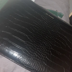 Svart datorfodral i croco-mönstrat skinn - Snygg svart kuvertväska från ideal of sweden i croco-mönstrat skinnimitation. Datorfodralet har silverfärgad dragkedja och en stilren, rektangulär form. Perfekt att bära under armen och rymmer både mobil och plånbok men också en dator . Mått ca 23x32 cm.