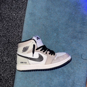 Nike Air Jordan 1 Gore-Tex vit/svart - Nike Air Jordan 1 Gore-Tex i vitt och grått med svarta detaljer och svart swoosh. Skorna har högt skaft, svarta snören och är tillverkade i en mix av skinn och syntetmaterial. Perfekt för dig som vill ha en ikonisk sneaker med vattentätt material.