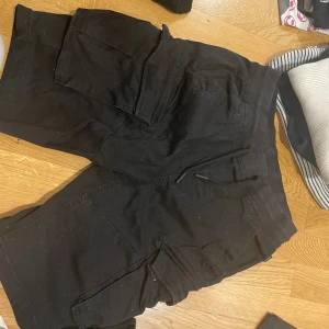 Svarta cargojorts från Cropp - Svarta cargoshorts från Cropp med flera stora fickor på sidorna och snörning i midjan. Regular fit och riktigt sköna för chill dagar. Perfekta för dig som gillar streetstyle och vill ha plats för allt i fickorna.