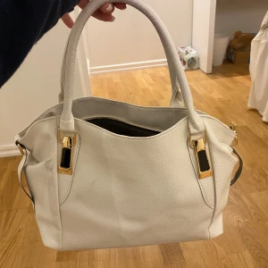 Vit handväska med gulddetaljer - Snygg vit handväska i skinnimitation med guldfärgade metalldetaljer och praktiska handtag. Väskan har en dragkedjeficka på baksidan och rymligt huvudfack med innerficka. Perfekt för dig som vill ha en stilren och klassisk look. De får plats en dator samt olika böcker och mycket mer. Helt ny oanvänd. Kan gå ner i pris vid snabb affär 