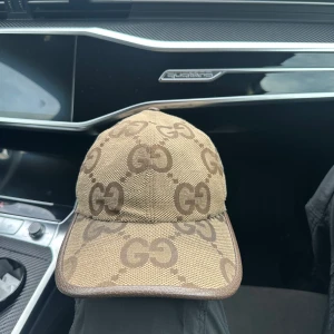 Gucci beige keps, Box o allt Kmr med - Snygg beige keps från Gucci med det klassiska GG-mönstret i brunt över hela kepsen. Kepsen har böjd skärm och justerbart band bak i brunt skinn. Materialet är en mix av bomull och skinn, perfekt för dig som gillar exklusiva accessoarer med tydlig designerstil.