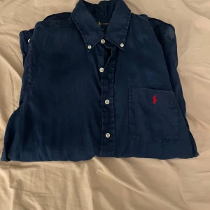 Mörkblå skjorta från Ralph Lauren - Klassisk mörkblå skjorta från Ralph Lauren i linne med vit knappstängning och röd broderad logga på bröstfickan. Skjortan har lång ärm, button down-krage och en tidlös, avslappnad passform.