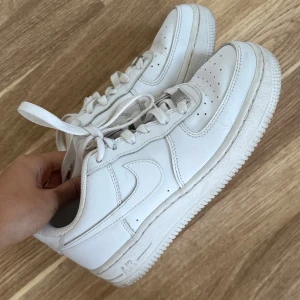 Vita Nike Air Force 1 sneakers - Klassiska helvita Nike Air Force 1 sneakers i skinn med rund tå, snörning och platt sula. Perfekta för dig som gillar en clean och tidlös look. Ikonisk modell med diskret Nike-logga på sidan. Storlek 36,5