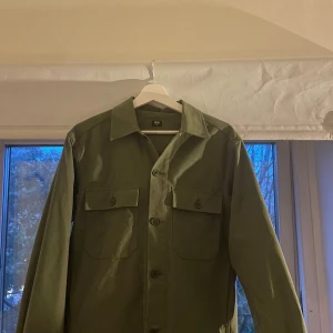  overshirt från Uniqlo - Stilren grön overshirt från Uniqlo med klassisk krage och två stora bröstfickor med knappar. Skjortan har raka linjer, lång ärm och stängs med knappar framtill. Perfekt lager-på-lager-plagg i bomull.