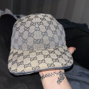 Intressekoll - Säljer en beige keps från Gucci med klassiskt GG-monogram i mörkblått över hela kepsen. Baktill finns en justerbar rem i mörkblått läder och en ikonisk röd-blå rand. Kepsen är tillverkad i bomullscanvas och har en böjd skärm.