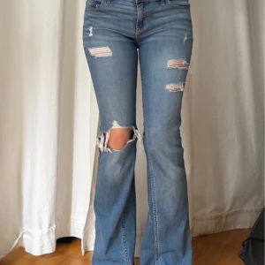 Blå bootcut jeans med slitningar - Snygga blå bootcut jeans med slitna detaljer och hål på knäna. Jeansen har låg midja och klassiska fickor bak med dekorativa sömmar. Perfekta för en avslappnad och trendig look. Midjemått är 40 cm rakt över och för referens är tjejen på bilderna 170 cm.