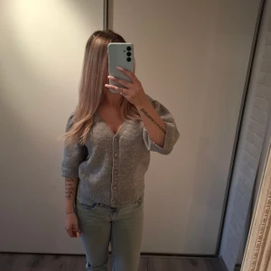 Grå kofta med puffärm och knappar - Superfin grå kofta med v-ringning och puffärmar. Koftan har knappar framtill och är stickad i ett mjukt material. Perfekt till jeans eller kjol för en chill och snygg look.