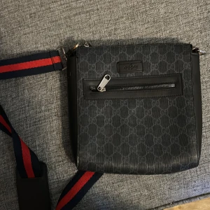 Svart axelväska från Gucci - Säljer en svart axelväska från Gucci med klassiskt GG-mönster och justerbar axelrem i marinblått och rött. Väskan har en dragkedjeficka framtill och detaljer i skinn. Perfekt för dig som vill ha en snygg och praktisk väska med lyxig känsla.