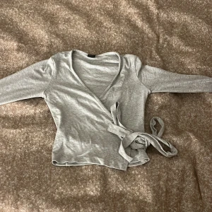 Grå omlottopp från Gina Tricot - Snygg grå omlottopp från Gina Tricot i mjuk bomullsmix. Modellen har långa ärmar, v-ringning och knyt i sidan för en cool och feminin look. Perfekt att styla med jeans eller kjol för en chill vibe.