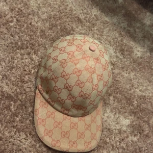 Beige och rosa Gucci keps - Snygg keps från Gucci i beige och rosa med klassiskt GG-mönster över hela kepsen. Justerbar rem bak i rosa skinn och liten guldfärgad GG-detalj på sidan. Materialet är bomull och skinn, perfekt för dig som vill sticka ut med lyxig streetstyle.
