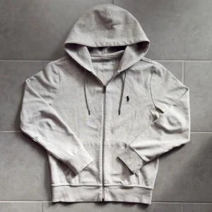 Grå hoodie från Polo Ralph Lauren - Snygg ljusgrå hoodie från Polo Ralph Lauren med dragkedja och huva. Tröjan har snörning i huvan, två fickor fram och den klassiska broderade loggan på bröstet. Tillverkad i mjuk bomull, perfekt för chill dagar och enkel att matcha med jeans.