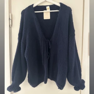 Mörkblå stickad kofta med knyt - Mysig mörkblå stickad kofta från TX Fashion med v-ringad hals och knyt framtill. Koftan har långa, puffiga ärmar med fin detalj vid handleden! Helt ny med prislapp kvar 