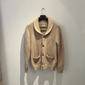 Beige stickad kofta från Made in the Shade - Säljer en beige stickad kofta från Made in the Shade i storlek M. Koftan har bred krage, ribbade muddar och två framfickor. Stängs med knappar framtill och har långärmade ärmar. Perfekt för lager-på-lager under kyliga dagar, den är i ull vilket gör den riktigt varm och mysig. Hör av er vid frågor