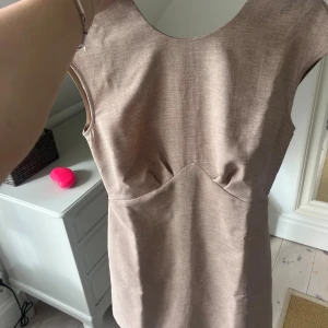 Beige ärmlös kortklänning med dragkedja - Snygg beige kortklänning med ärmlös design och markerad midja. Klänningen har en stilren look med diskret struktur i tyget och en guldig dragkedja i ryggen som detalj. Perfekt för dig som gillar minimalistisk och clean stil.
