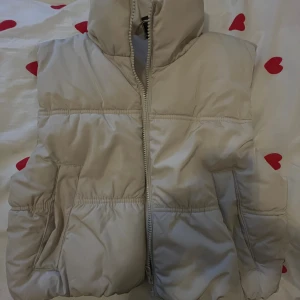 Beige dunväst från H&M Divided S - Beige dunväst från H&M Divided i storlek S. Västen har hög krage, dragkedja framtill och två fickor på sidorna. Den är quiltad och har en puffig look som ger en trendig och avslappnad vibe. Perfekt att slänga över en hoodie eller tröja.