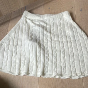 Vit stickad kortkjol från SisterX - Supersöt vit stickad kortkjol från SisterX med kabelstickat mönster och bred resår i midjan. Kjolen har en mjuk och mysig känsla, perfekt för att skapa en preppy look. Färgen är star white och modellen är lätt utställd.