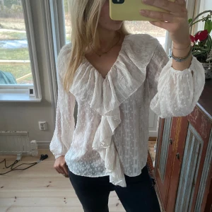 Vit blus med volang från MNG - Säljer en vit blus från MNG i storlek M. Blusen har ett snyggt volangparti längs v-ringningen och framtill, samt diskret prickigt mönster i tyget. Ärmarna är långa och lätt puffiga med resår vid mudden. Materialet är tunt och luftigt, perfekt för vår, sommar men också finare tillfällen året om😍