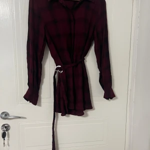 Vinröd rutig skjortklänning med bälte - Vinröd och svart rutig skjortklänning med långa ärmar och knappar vid ärmslut. Klänningen har ett avtagbart bälte i midjan med metallspänne och klassisk krage. Mjuk och lätt bomullskänsla, perfekt för en chill och snygg look. Den har vissa färgfläckar och även eventuellt små hål.