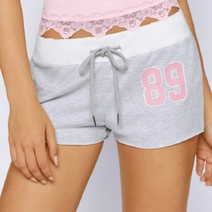 Grå shorts med snörning och tryck - Snygga grå shorts med vit midja och snörning framtill. På ena benet finns ett rosa tryck med siffran 89. Perfekta för chill dagar eller träning. Mjukt material som känns skönt mot huden och ger en avslappnad vibe.