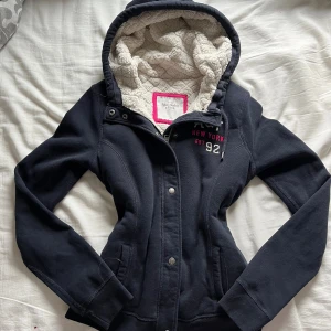 Marinblå hoodiejacka från Abercrombie & Fitch - Mysig marinblå hoodiejacka från Abercrombie & Fitch med broderad logga och text på bröstet. Jackan har vit teddyfoder i huvan, dragkedja och tryckknappar framtill samt två fickor. Perfekt för kyliga dagar och har en sportig, avslappnad vibe.