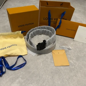 Louis Vuitton svart skinnbälte - Snyggt Louis Vuitton bälte i svart skinn med klassisk LV-logga i svart metall på spännet. Bältet har ett diskret rutigt mönster och kommer med originalförpackning, dustbag och blått band. Perfekt accessoar för att lyfta din outfit.