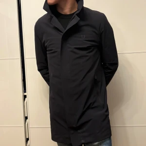 Marinblå rock från UBR med dragkedja - Snygg marin blå rock från UBR med stilren design och hög krage. Jackan har diskreta fickor med dragkedja och ett minimalistiskt UBR-logo på bröstet. Perfekt längd och klassisk passform, tillverkad i ett slätt syntetmaterial som står emot väder och vind.