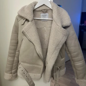 Beige pilotjacka från Stradivarius - Säljer en snygg beige pilotjacka från Stradivarius med bred krage, dragkedja och bälte i midjan. Jackan har mjukt teddyfoder och detaljer vid ärmslut och nederkant. Perfekt för kalla dagar. Köpte denna förra vintern men den kom inte till användningen mer än 5 gånger. 