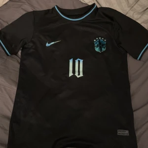 Svart Brasilien Neymar Jr Nike tröja - Säljer en svart Brasilien fotbollströja från Nike med Neymar Jr och nummer 10 på ryggen. Tröjan har korta ärmar, blå detaljer vid krage och ärmslut samt Brasilien-emblem med fem stjärnor. Tillverkad i Dri-FIT material som andas och håller dig torr. Storlek M