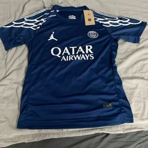 PSG x Jordan blå fotbollströja - Blå PSG x Jordan fotbollströja med vita detaljer och tryck. Kortärmad med unikt mönster på axlarna, Qatar Airways-logga på bröstet och Jordan-logga. Tillverkad i Dri-Fit material som andas, perfekt för träning eller match.