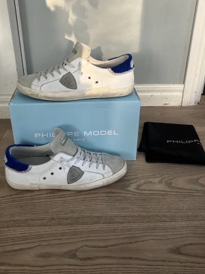 Philippe Model vita sneakers med blå detalj - Snygga vita Philippe Model sneakers med grå mockadetaljer och blå fluffig häl. Klassisk låg modell med vita skosnören och gröna logotaggar på plösen. Skorna har en ikonisk sköld på sidan och slitstark gummisula. Perfekta för en trendig streetstyle-look. Har kvitto på skorna. Användna i bara 2 månader och är mycket bra skick. Inte mycket prutning för vi har redan sänkt riktigt mycket.👍