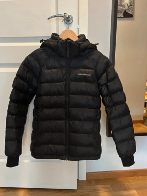 Svart dunjacka från Peak Performance - Snygg svart dunjacka från Peak Performance med huva och dragkedja framtill. Jackan har diskret logga på bröstet, ribbade muddar vid ärmsluten och två sidofickor. Perfekt för kalla dagar och riktigt varm tack vare dunfyllningen. Storlek 160