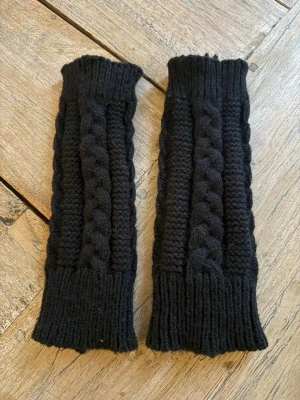 Svarta stickade pulsvärmare - Ett par svarta stickade pulsvärmare med flätmönster. De är långa och har öppning för tummen, vilket gör dem perfekta för att hålla händerna varma samtidigt som fingrarna är fria. Passar perfekt till kyliga dagar och ger en mysig touch till din outfit. Oanvända.