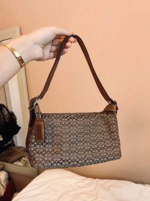 Brun axelväska från Coach - Säljer en brun axelväska från Coach med det klassiska C-mönstret över hela väskan. Väskan har en justerbar axelrem i brunt läder och en praktisk ytterficka med dragkedja på baksidan. Perfekt storlek 