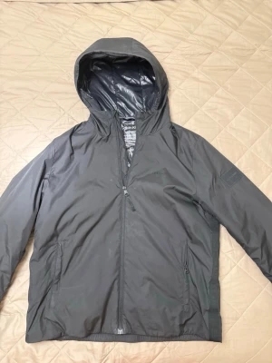 Svart vindjacka från Calvin Klein - Snygg svart vindjacka från Calvin Klein med huva och dragkedja framtill. Jackan har två sidofickor med dragkedja och är tillverkad av återvunnen polyester. Perfekt för blåsiga dagar och enkel att matcha med olika outfits.