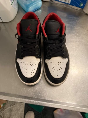 Svarta och vita Air Jordan 1 Low - Säljer ett par Air Jordan 1 Low sneakers i svart och vitt med röda detaljer och röd insida. Skorna har klassisk perforerad tåbox, svarta skosnören och Jumpman-logga på plösen. Tillverkade i läder och textil för en clean och sportig look.