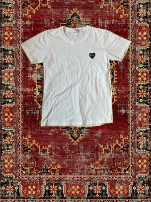 Vit t-shirt från Comme des Garçons Play - Vit t-shirt från Comme des Garçons Play med det ikoniska svarta hjärtat på bröstet. Klassisk passform och rund halsringning. Tillverkad i mjuk bomull som känns skön mot huden. Perfekt för en clean och stilren look.
