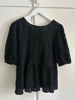 Svart strukturerad blus med puffärm - Säljer en svart blus med puffiga korta ärmar och vågig struktur i tyget. Blusen har en lös passform, volang nedtill och en snygg knytning i ryggen. Perfekt till jeans eller kjol för en trendig look.