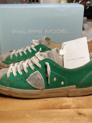 Gröna Philippe Model sneakers - Snygga gröna sneakers från Philippe Model med silverdetaljer och vita skosnören. Skorna har en låg profil, vit sula och en distinkt logotyp på sidan. Ovandelen är i läder med en vintagekänsla och insidan är mjukt fodrad för extra komfort.