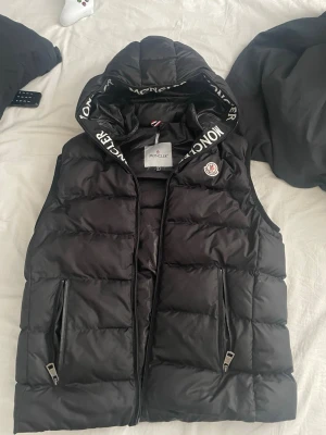 Svart dunväst från Moncler med huva - Snygg svart dunväst från Moncler med avtagbar huva och dragkedjor på fickorna. Västen har Moncler-logga på bröstet och textdetaljer på huvan. Perfekt för lager-på-lager och streetstyle. Tillverkad i glansigt material med klassisk puffer-look. Har ett hål i kragen som är enkelt och sy, men eftersom jag inte orkar göra det säljer jag den. Hålet syns inte när du har den på dig. 