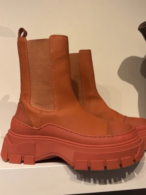 Chunky orange Chelsea boots från MNG - Säljer ett par chunky Chelsea boots i orange från MNG. Boots i slätt konstläder med bred resår på sidorna och grov platåsula. Högt skaft och dragflik bak. Perfekta för dig som vill sticka ut med färg och cool siluett.