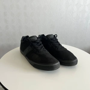 Svarta Polo Ralph Lauren sneakers - Snygga helsvarta sneakers från Polo Ralph Lauren med lågt skaft. Skorna har en klassisk siluett, ovandel i textil och detaljer i läderimitation. Märkets logga syns på sidan och tungan. Perfekta för en stilren och avslappnad look.