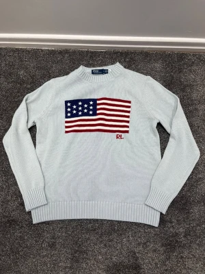 Ljusblå stickad tröja Polo Ralph Lauren - Ljusblå stickad tröja från Polo Ralph Lauren med amerikansk flagga i rött, vitt och blått på bröstet samt RL-broderi. Tröjan har rund hals, ribbade muddar och nederkant. Perfekt för dig som gillar klassisk preppy stil.
