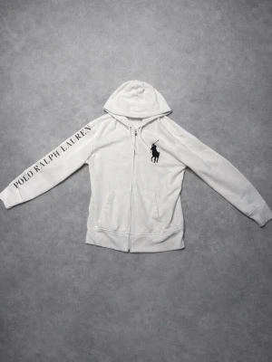 Vit hoodie från Polo Ralph Lauren - Snygg vit hoodie med dragkedja från Polo Ralph Lauren. Stor logga med polospelare på bröstet och texten 'POLO RALPH LAUREN' längs ena ärmen. Mjuk bomullskänsla, två fickor fram och klassisk huva. Perfekt för en clean och sportig look.
