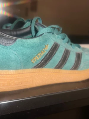 Adidas Spezial mocka - Säljer ett par Adidas Spezial sneakers i turkos mocka med svarta läderdetaljer och klassiska tre ränder på sidan. Skon har guldfärgade loggor och en gummisula i beige. Perfekt för dig som gillar retrostil och vill sticka ut.