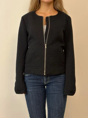 Svart quiltad cardiganjacka med dragkedja - Svart cardiganjacka med quiltat sicksackmönster och silverfärgad dragkedja framtill. Jackan har rund halsringning och långa ärmar. Materialet ser mjukt och lite stretchigt ut, perfekt för lager-på-lager-stil.