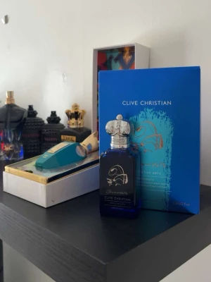 Clive Christian blå parfymflaska - Lyxig parfym från Clive Christian i en djupblå glasflaska med silverfärgad, detaljerad kork. Flaskan har en elegant design med guldmönster och levereras i en matchande blå förpackning med exklusiv känsla. Perfekt för dig som gillar unika och exklusiva dofter.